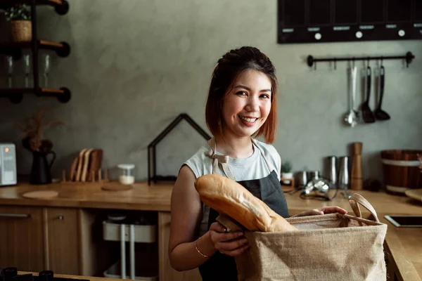 Asian woman buying couple of french bread, putting them in cloth paper bag.