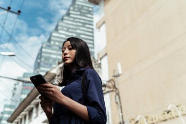 Mavi elbiseli güzel Asyalı kadın şehirde ona rehberlik etmek için akıllı telefondan GPS kullanıyor..