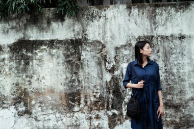 Mavi elbiseli, siyah çantalı, kirli duvarda duran güzel Asyalı kadın..