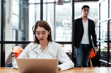 Kulaklık takan, bilgisayarda mikrofon takan çağrı merkezi kadını müşteriyle konuşurken patronu da nasıl çalıştığını görmek için kapıyı dikizliyor..