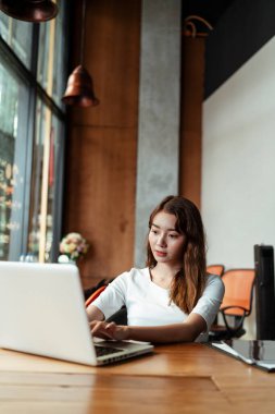 Beyaz elbiseli Asyalı genç kadın modern ofis odasında dizüstü bilgisayarla çalışmaya odaklanmış..