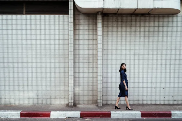 Mavi elbiseli güzel Asyalı kadın beyaz duvardaki kaldırımda yürüyor..