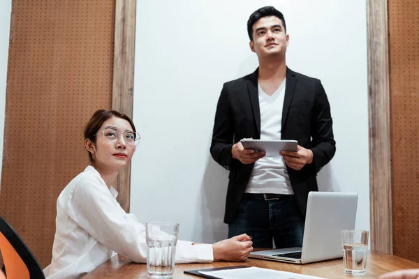 Asian business boss in black suit holding laptop showing presentation on the screen to show how the company work to young asian lady on white shirt wearing eyeglasses sitting in the room.