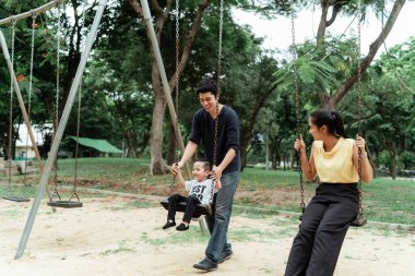 Küçük bir çocuk annesinin yanında salıncakta otururken babası onu nazikçe sırtından tutuyor, hey birlikte gülün..