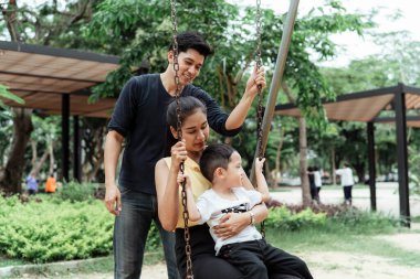 Küçük çocuk annesinin kucağında otururken kız parkta salıncakta oturuyor ve babası da arkada ona destek oluyor..