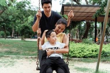 Küçük çocuk annesinin kucağında otururken kız parkta salıncakta oturuyor ve babası da arkada ona destek oluyor..