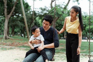 Baba salıncakta oturuyor ve oğlu kucağında oturuyor, anne onların yanında duruyor, ailenin en güzel zamanları..
