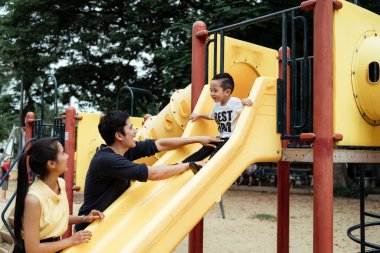 Anne ve baba, parkta mutlulukla kaydırma oynayan oğullarına destek olmak için yerde duruyorlar..