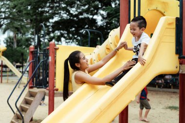 Annesi oğlunu beklerken sarı kaydıraktan aşağı kayıyor, ikisi de mutlu bir şekilde gülümsüyor..