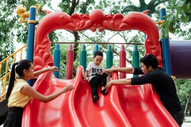 Beyaz tişörtlü küçük çocuk, ailesi onunla ilgilenirken kırmızı oyun parkı kaydırma oyunu oynuyor..