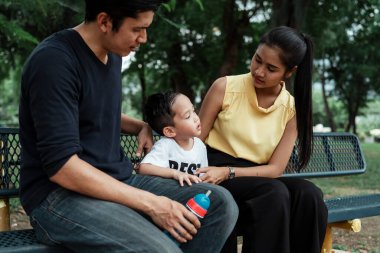 Küçük çocuk ailesiyle bankta oturuyor ve onlarla konuşuyor..