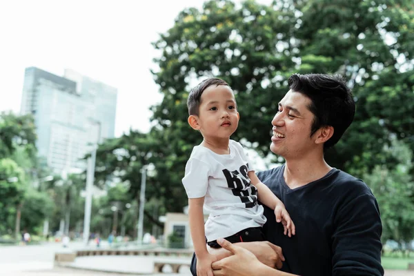 Siyah tişörtlü bekar bir baba hafta sonu parkta seyahat ederken oğlunu taşıyor..