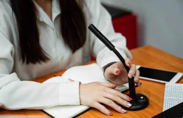 Black microphone is brabbed by the woman with nail painting on the wooden table with smartphone and notebook.
