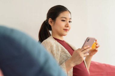 Güzel hamile Asyalı kadın portakal suyu tutuyor ve akıllı telefon kullanıyor. Kanepede oturuyor..