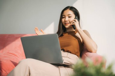 Güzel Asyalı Taylandlı iş kadını dizüstü bilgisayar ve akıllı telefonla çalışıyor, evdeki bir kanepede oturuyor. Evden çalış..