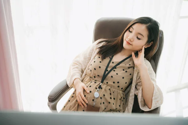 Beautiful pregnant asian thai businesswoman resting on a chair and holding belly at office room.