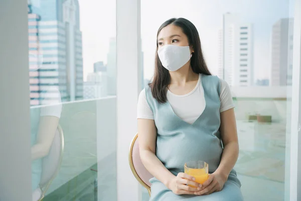 オレンジ色のジュースのガラスを保持する椅子に座って綿マスクを着て美しい妊娠中のアジアの女性 自宅での隔離 — ストック写真