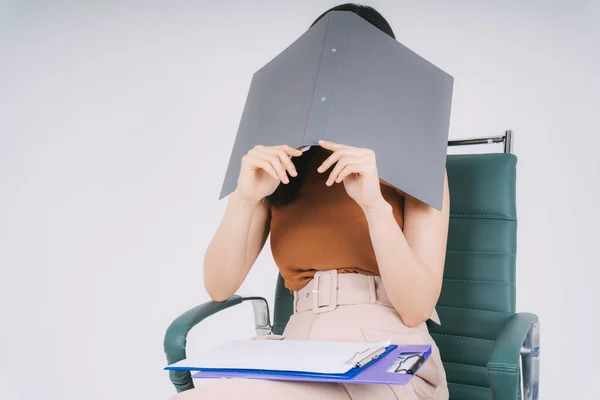 Beautiful asian thai businesswoman sitting on a chair covering her face with a big folder document. Isolation over white background.