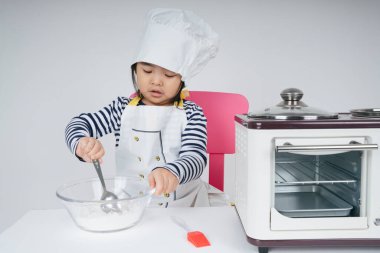 Tatlı Asyalı Taylandlı çocuk mutfakta un ve suyu cam kaseye karıştırarak şef rolünde oynuyor..