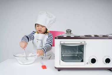 Tatlı Asyalı Taylandlı çocuk mutfakta un ve suyu cam kaseye karıştırarak şef rolünde oynuyor..