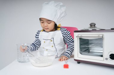 Tatlı Asyalı Taylandlı çocuk mutfakta un ve suyu cam kaseye karıştırarak şef rolünde oynuyor..