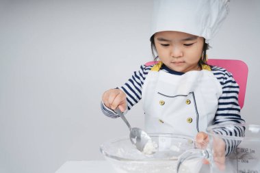 Tatlı Asyalı Taylandlı çocuk mutfakta un ve suyu cam kaseye karıştırarak şef rolünde oynuyor..