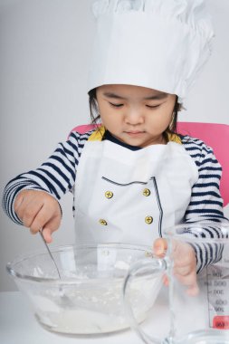 Tatlı Asyalı Taylandlı çocuk mutfakta un ve suyu cam kaseye karıştırarak şef rolünde oynuyor..