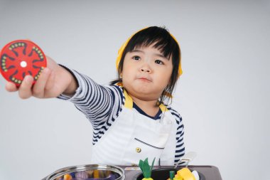 Güzel Asyalı Taylandlı çocuk, mutfakta elinde domates dilimi tutan sarı bandajlı bir şef rolünde oynuyor..