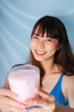 Neşeli genç Asyalı siyah saçlı kadın iki eliyle bir bardak pembe milkshake tutuyor..