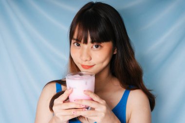 Neşeli genç Asyalı siyah saçlı kadın iki eliyle bir bardak pembe milkshake tutuyor..