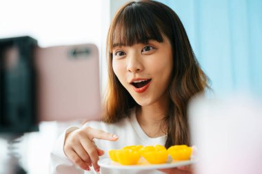 Genç Asyalı siyah saçlı kadın akıllı telefon kamerasının önünde Tayland tatlısı incelemesi yapıyor..