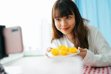 Genç Asyalı siyah saçlı kadın akıllı telefon kamerasının önünde Tayland tatlısı incelemesi yapıyor..