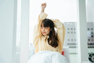 Genç Asyalı siyah saçlı kadının portresi sabah yatakta uyanır. Kollarını açan kız uyandıktan sonra rahatlar..