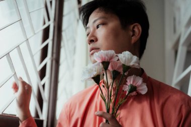 Turuncu mandalina yakalı eşcinsel adam pencerenin yanında oturuyor çiti tutuyor ve dışarı bakıyor pembe ve beyaz karanfili nazikçe tutarken.