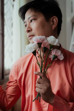 Turuncu mandalina yakalı eşcinsel adam pencerenin yanında oturuyor çiti tutuyor ve dışarı bakıyor pembe ve beyaz karanfili nazikçe tutarken.