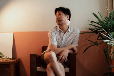 Soyunmuş tişörtlü bir eşcinsel sandalyede otururken karanfil çiçeğini ısırır..