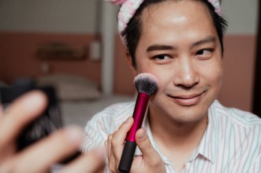 Asyalı eşcinsel makyajını tamamlamak için yanağını fırçalıyor..