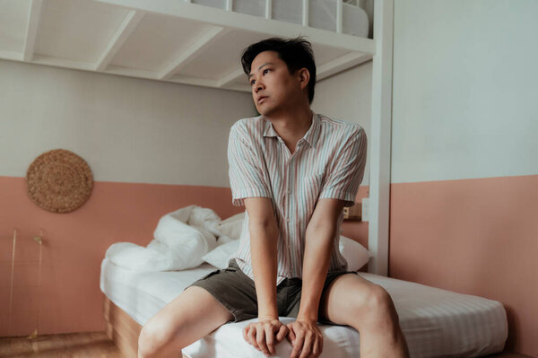 Asian gay in stripped shirt sitting on the white bed looking outside the window.