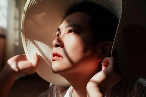 Asian gay chin up and face to the sun while he sitting at the window with white hat.