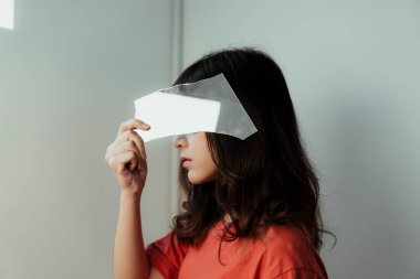 Turuncu tişörtlü kıvırcık saçlı kız yüzünü kırık aynayla örtüyor..