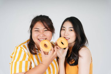 Sağlıksız çörek tutan iki Asyalı tombul ve zayıf kadın beyaz arka planda izole edilmiş..