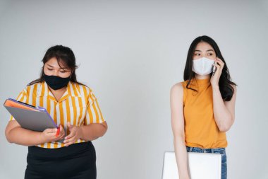 İki Asyalı iş kadını Covid-19 'u önlemek için maske takıyor. Tombul kadın dosya klasörünü tutuyor. Sıska kadın telefonda konuşuyor..