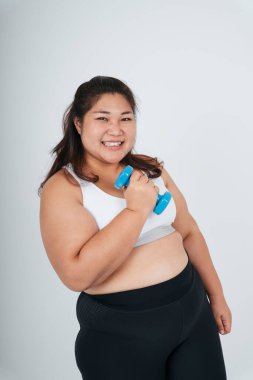 Beyaz spor sütyenli neşeli Asyalı tombul kadın beyaz arka planda mavi dambıl tutuyor..