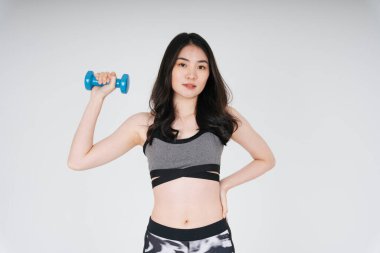 Beyaz arka planda dambılı gri spor sutyen egzersizi yapan güzel, ince Asyalı kadın..