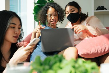 Çeşitlilik toplumu konsepti. Asyalı ve Afrikalı kadınlar evde dizüstü bilgisayar kullanarak yüz maskesi takıyorlar..