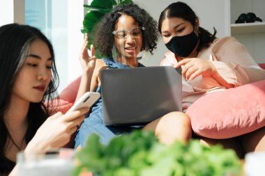 Çeşitlilik toplumu konsepti. Asyalı ve Afrikalı kadınlar evde dizüstü bilgisayar kullanarak yüz maskesi takıyorlar..