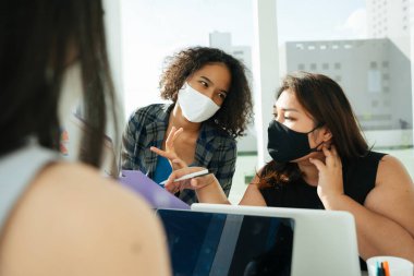 Üç farklı Asyalı ve Afrikalı iş kadınından oluşan bir grup ofiste birlikte çalışırken maske takıyor..