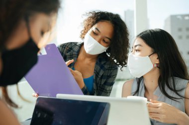 Üç farklı Asyalı ve Afrikalı iş kadınından oluşan bir grup ofiste birlikte çalışırken maske takıyor..