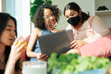 Afrikalı ve Taylandlı kadınlar dizüstü bilgisayar kullanarak yüz koruyucusu ve maske takarlar. Ev kavramından çalış.