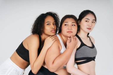 Beyaz arka planda spor sutyen giyen Asyalı ve Afrikalı kadınların grup portresi.